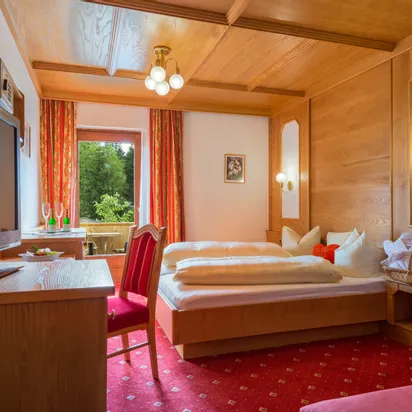 Gemütliches Hotelzimmer mit Holzmöbeln, Doppelbett, rotem Teppich und Balkonblick ins Grüne